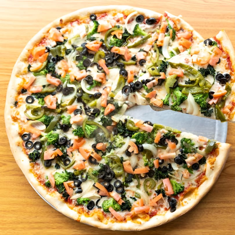 A large, whole pizza with a slice being lifted, topped with broccoli, olives, tomatoes, and peppers.