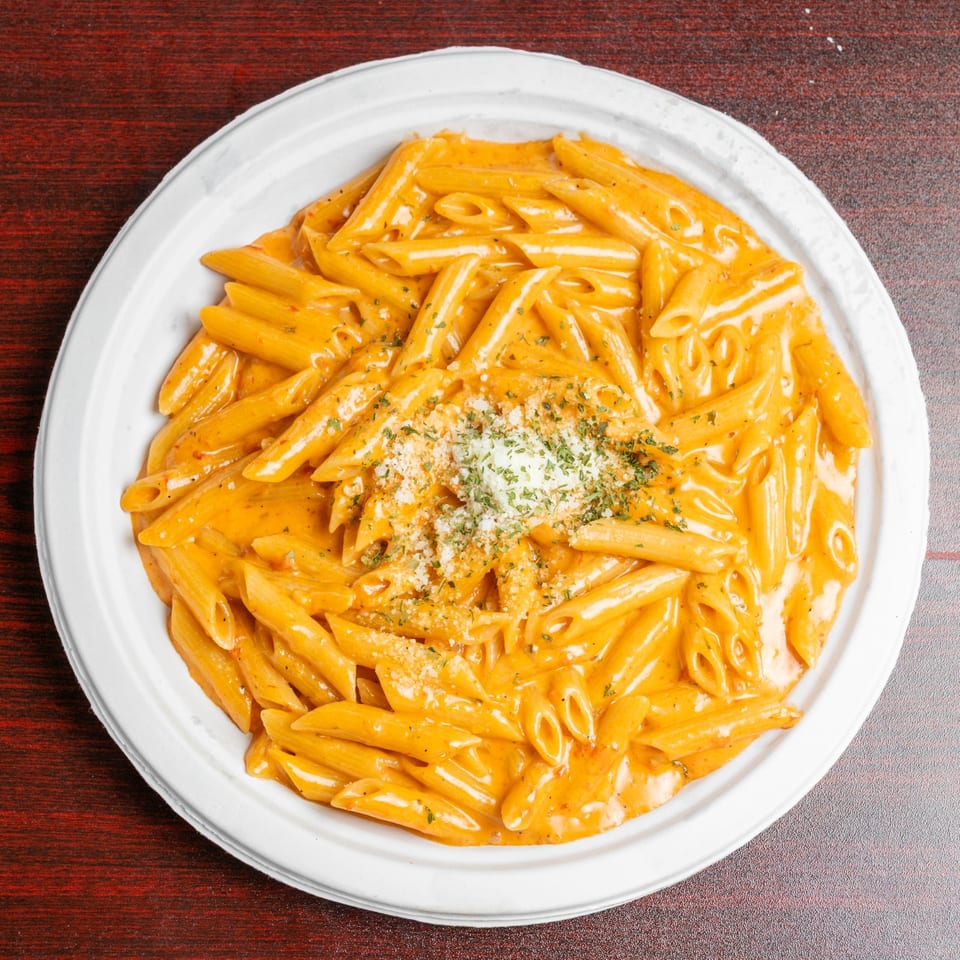 Penne pasta coated in creamy orange sauce, topped with grated cheese and herbs on a white plate.