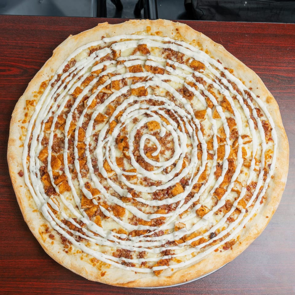 Large round pizza topped with browned meat, cheese, chicken pieces, and white sauce drizzled in concentric circles on a wooden table.