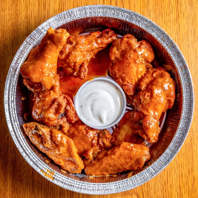A foil container filled with saucy buffalo chicken wings and a small cup of ranch dressing.
