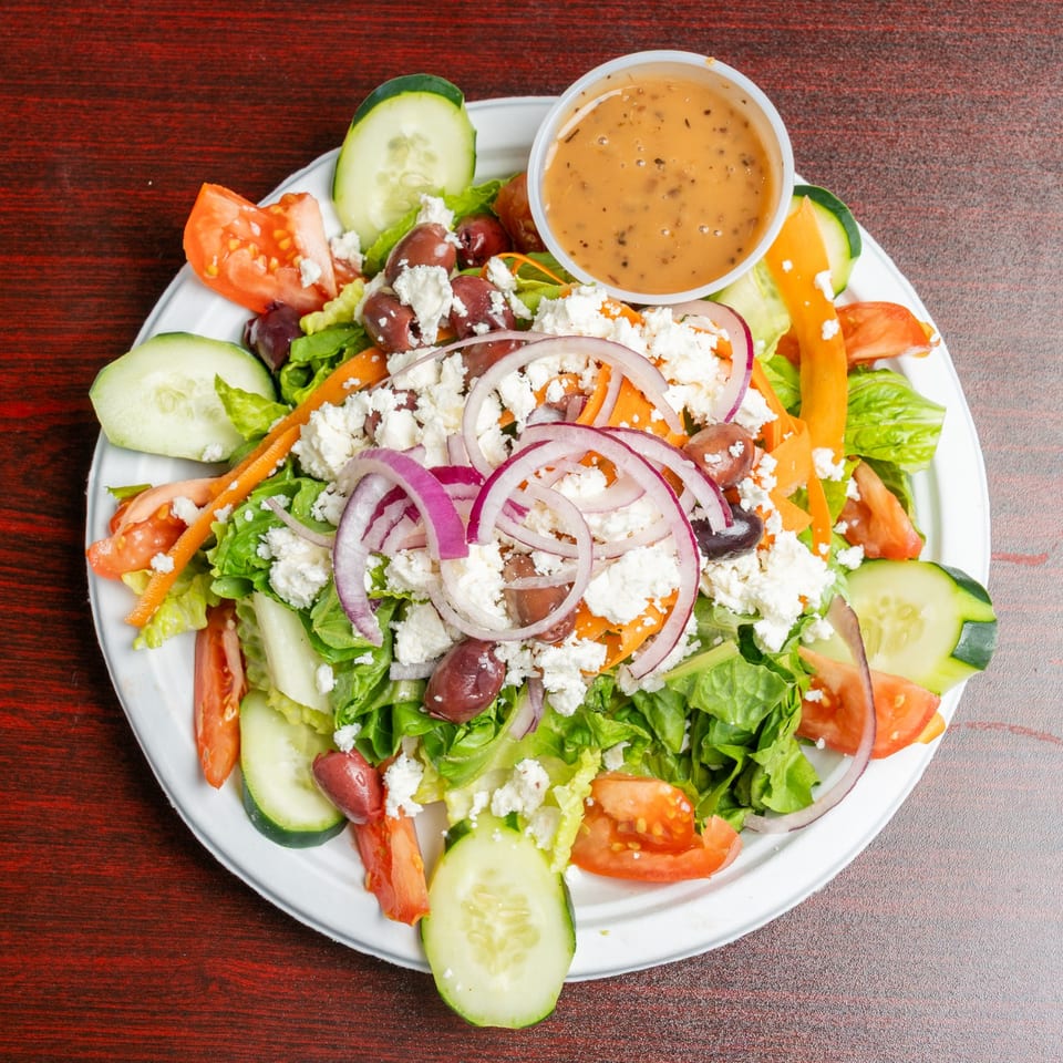 Greek salad with lettuce, cucumbers, tomatoes, olives, red onions, carrots, feta cheese, and dressing on a white plate.