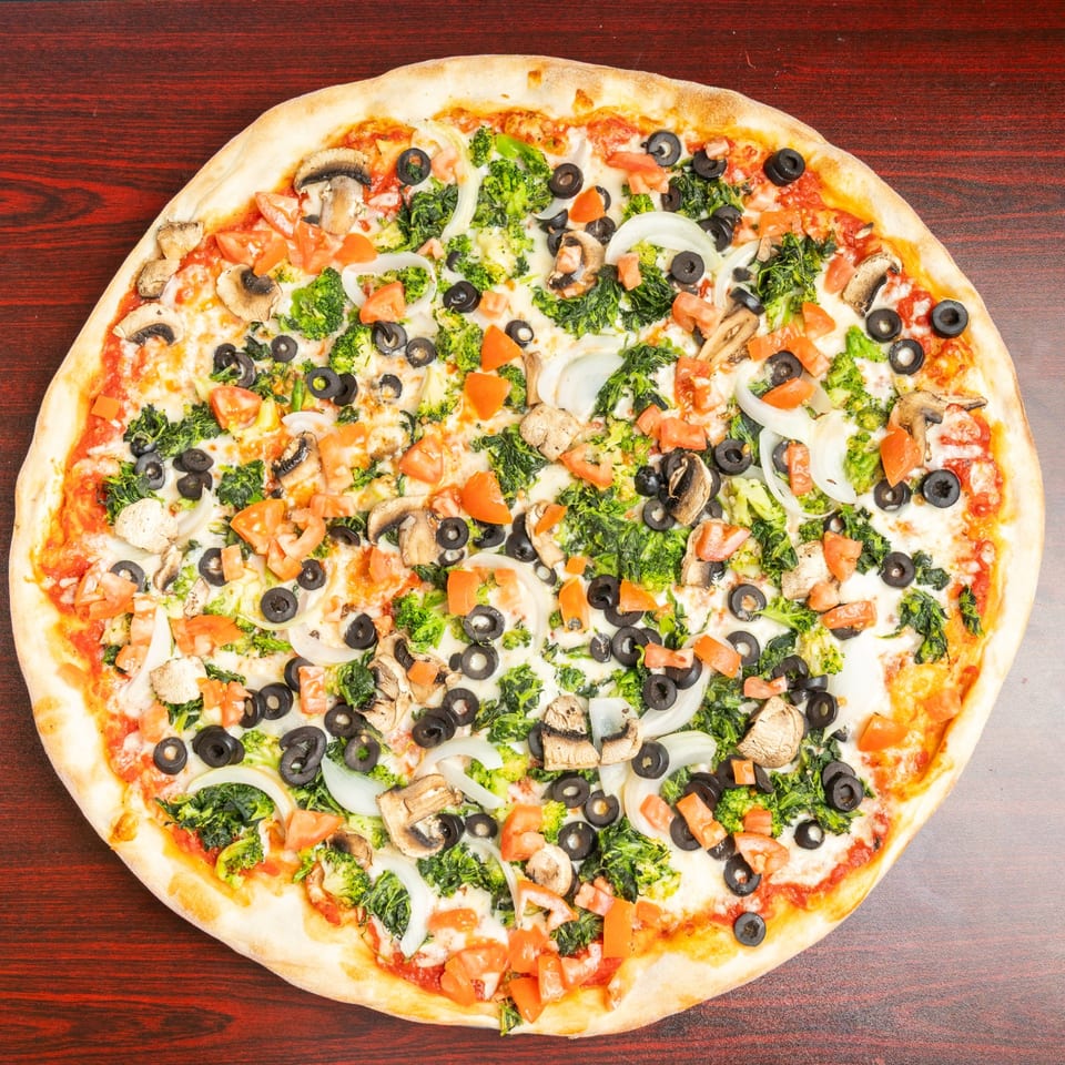Large vegetarian pizza topped with broccoli, spinach, mushrooms, black olives, tomatoes, onions, and melted cheese on a wooden table.
