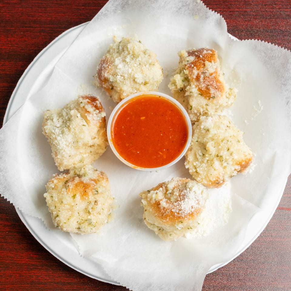 Five breaded, golden cheese bites arranged around a cup of bright red dipping sauce on a paper-lined plate.