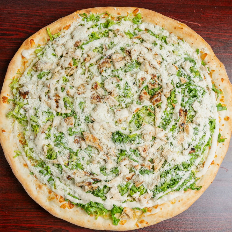 Round flatbread topped with chopped grilled chicken, lettuce, creamy dressing, and grated cheese on a dark wooden table.