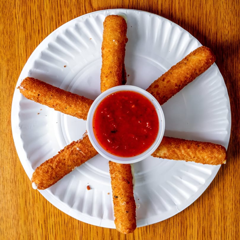 Golden-brown mozzarella sticks arranged around a cup of red marinara sauce on a white paper plate.
